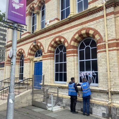 Two clean team workers, wearing blue vests, clean off graffiti from a window vinyl on the Shaftesbury Hospital.