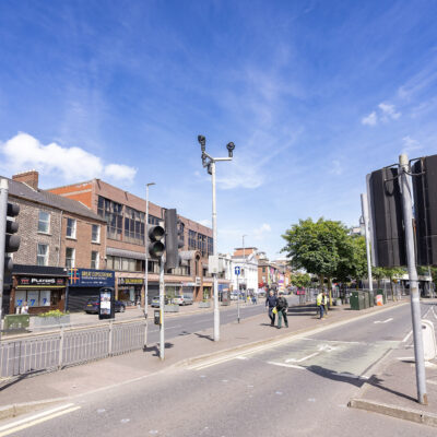 This image captures a sunny day on Shaftsbury Square in Belfast. The sky is very blue, and there are some people walking on the street.