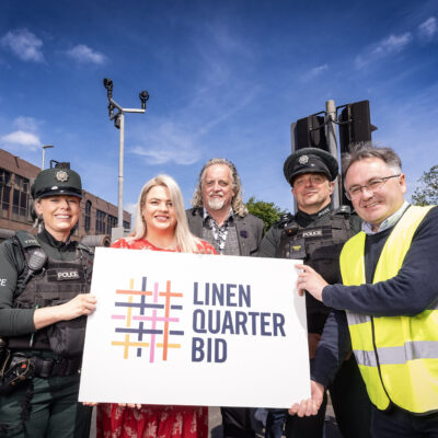 This image shows two police officers, the Belfast Night Czar, a Linen Quarter Employee, and a member of the Clean Team standing in a line smiling at the camera, holding a while Linen Quarter BID sign towards the camera. The day is sunny with a blue sky.