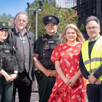 This image shows two police officers, the Belfast Night Czar, a Linen Quarter Employee, and a member of the Clean Team standing in a line smiling at the camera. The day is sunny with a blue sky.