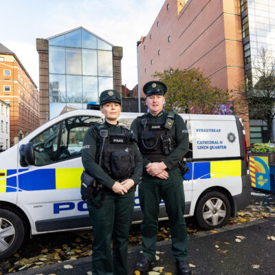 Two police officers (one male and one female) stand in front of a police van.