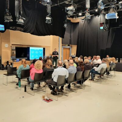 This image shows a group of people sitting in rows of chairs facing a police officer who stands at the front of the room and is giving a presentation.