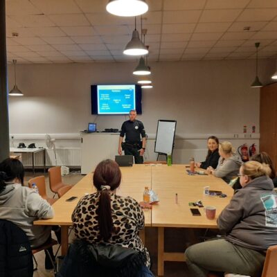 This image shows a group of woman sitting around a large table, with a police officer standing at the front of the room giving a presentation.