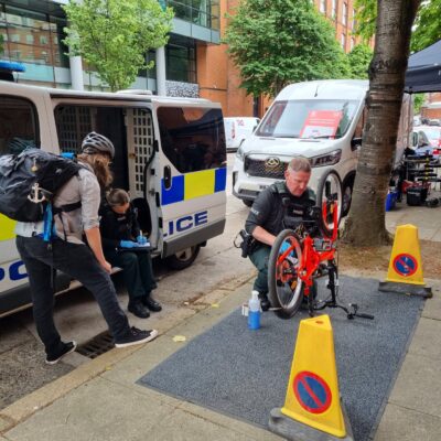 This image shows one police officer installing a device to a red overturned bike on the street. The bike owner stands to the side talking to an officer who is filling a form out. This activity being shown is from bicycle marking to promote anti-theft.