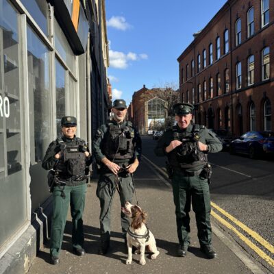 This image shows three police officers standing in a line on the footpath. One of the officers has a police dog with him, and they are all smiling for the camera. The day in Belfast is very sunny with a blue sky.