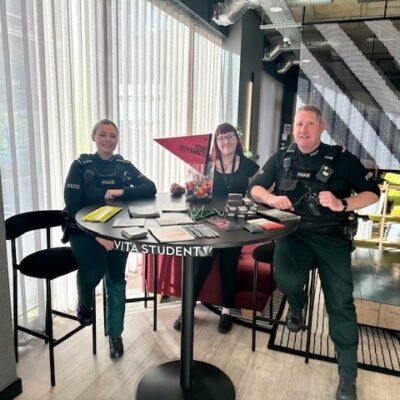 This image shows two police officers, sitting next to a woman at a table for a Vita Student event. They are smiling and sitting at a high table with brochures on it.