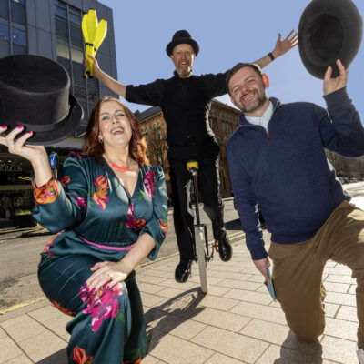 LQ BID's Stevie and Imagine Festivals Mags kneel on one knee, holding top hats, while a circus performer appears in the background on a unicycle holding juggling skittles