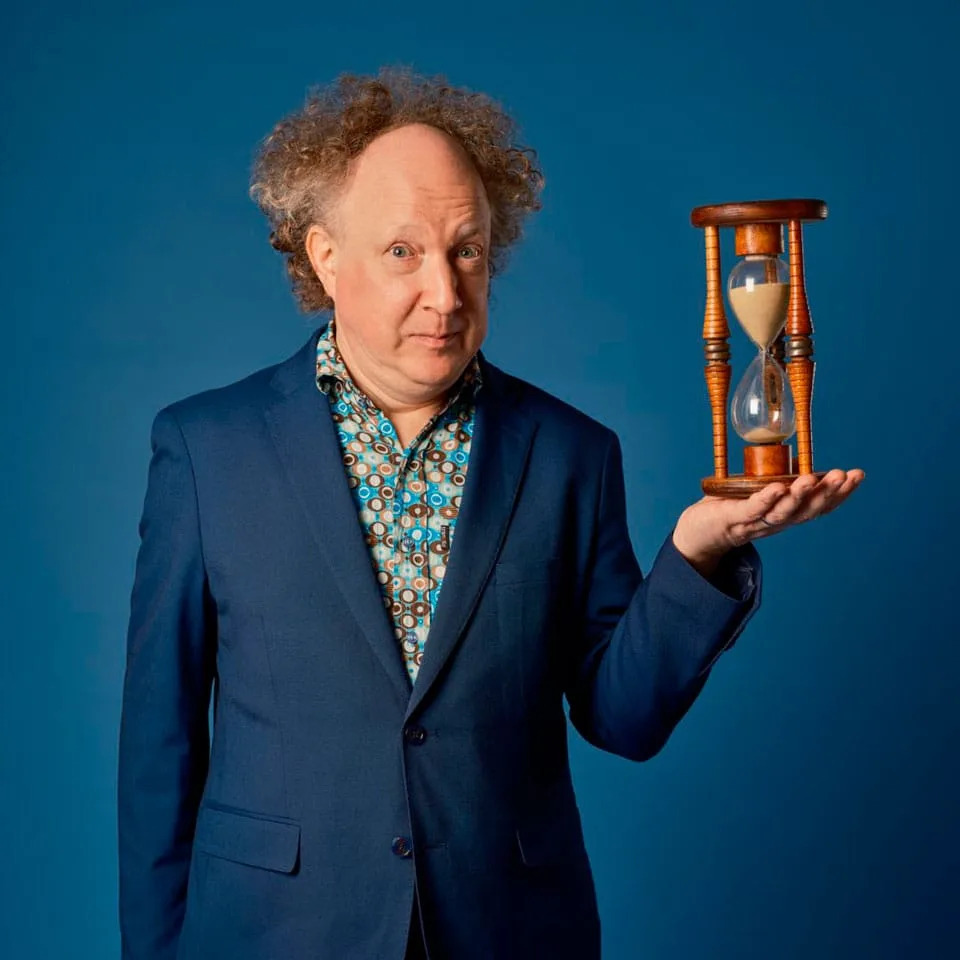 Andy Zaltzman in a blue suit holding an hourglass