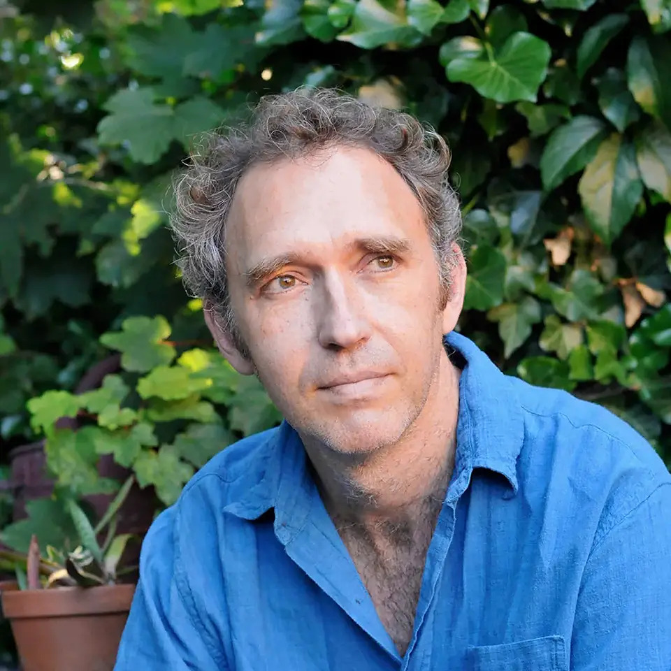 Picture shows Roman Krznaric in a blue shirt with a green bush in the background and a brown pot