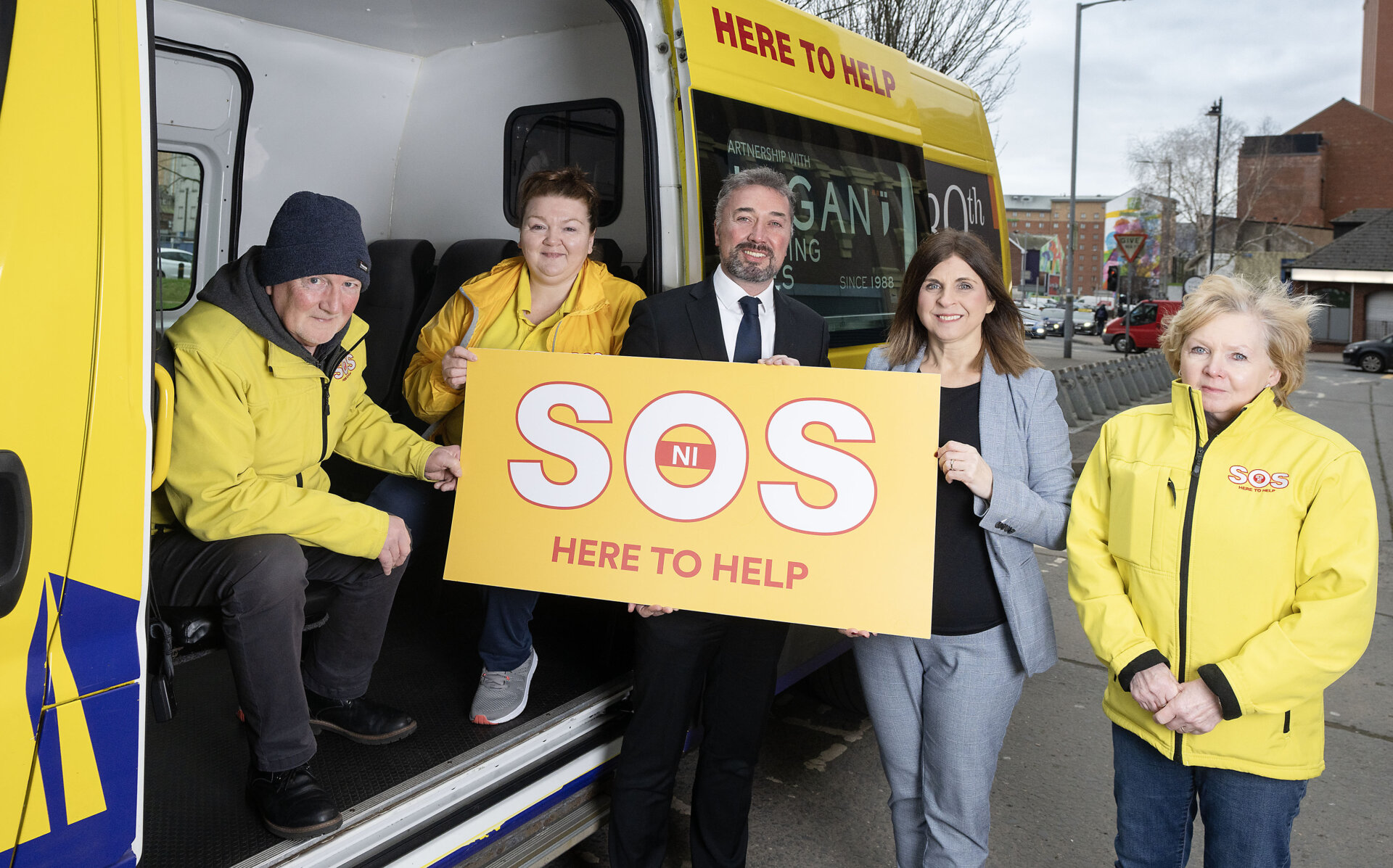 This picture shows SOS Bus workers in yellow jackets next to the Linen Quarter BID's Managing Director all holding a yellow SOS sign that reads, 