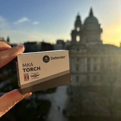 A hand holds a boxed safety alarm, with a view of Belfast City Hall as the background with the sunsetting.