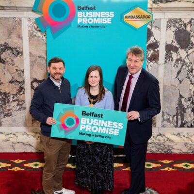 LQ Comms Manager Stevie Maginn poses with the High Sheriff of Belfast, Cllr Jenna Maghie, and the DfE Permanent Secretary. They are holding a Belfast Business Promise logo with 'ambassador' written in gold. The marble walls and windows appear in the background