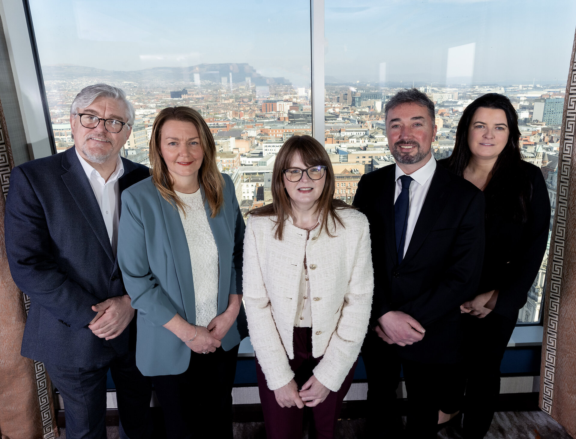 Three Belfast BID Managers, the Minister for the Economy and a representative from Tourism NI stand on the 23rd floor Observatory Bar at a window, with Belfast and Cave Hill in the background