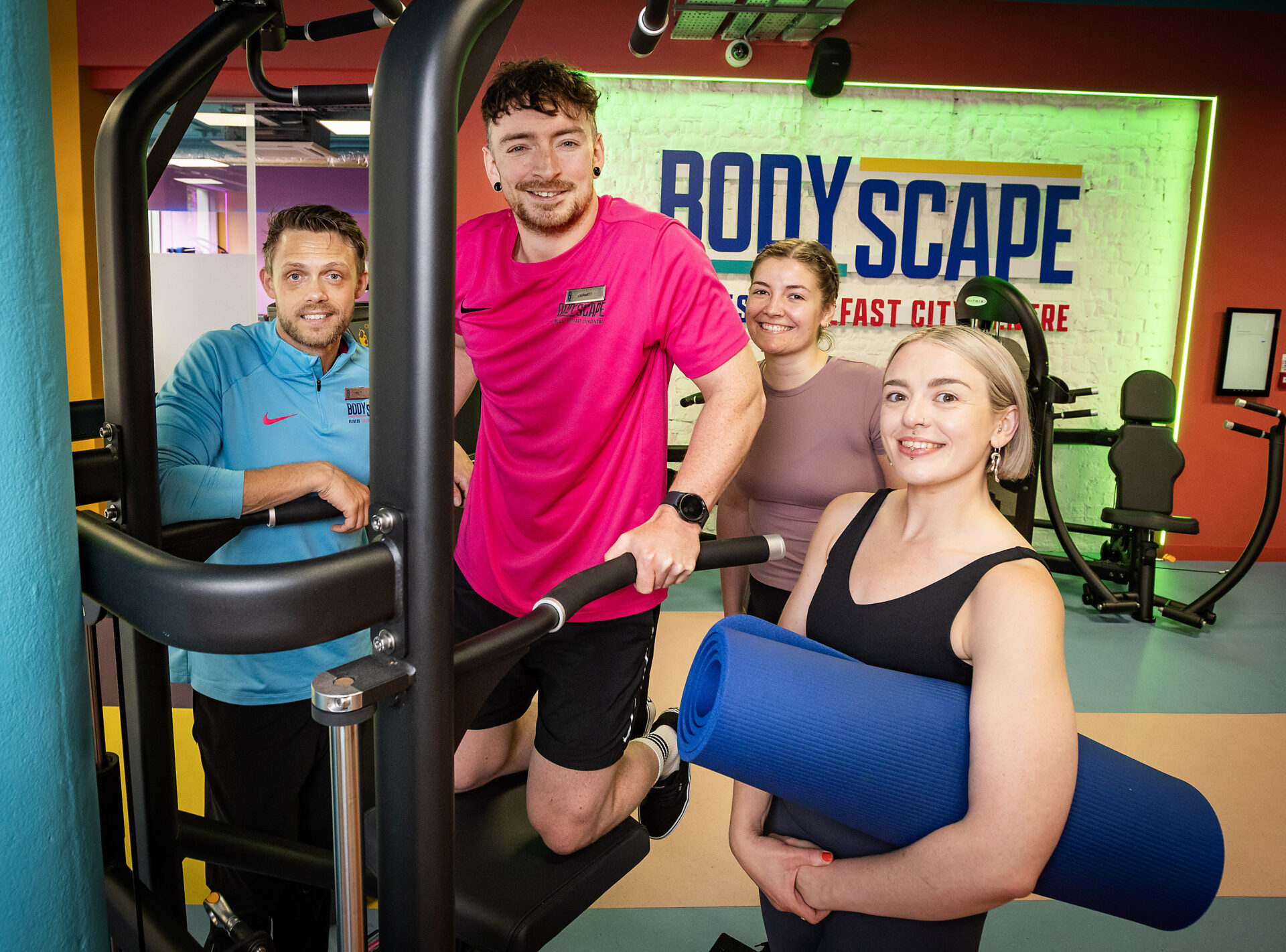 3 women and a man pose in a gym, one of the pen wears a pink top and is carrying out an assisted tricep excercise while a woman in front holds a blue yoga matt