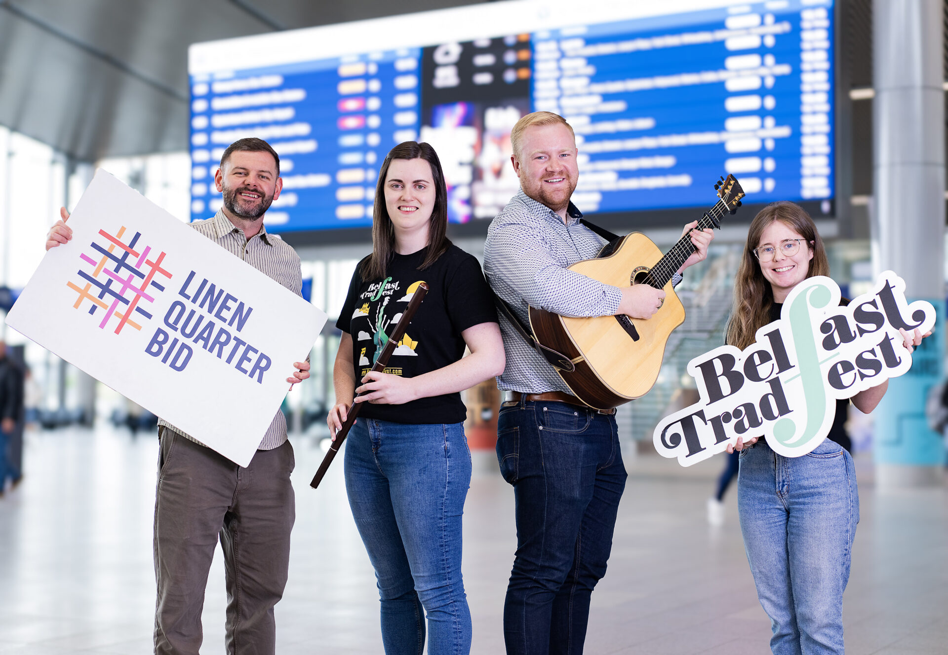 Two musicians, one female and one male, pose in a station holding a flute and a guitar, they are flanked by another man and woman holding foam boards with logos saying ; Belfast Tradfest & Linen Quarter BID'