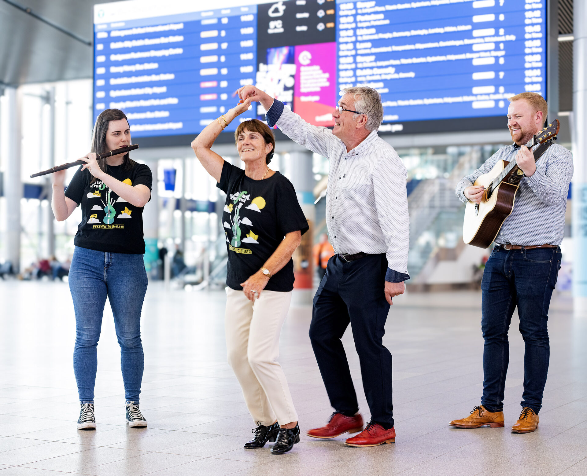 A woman plays flute while a man plays guitar in a station, while another man and woman dance inbetween them