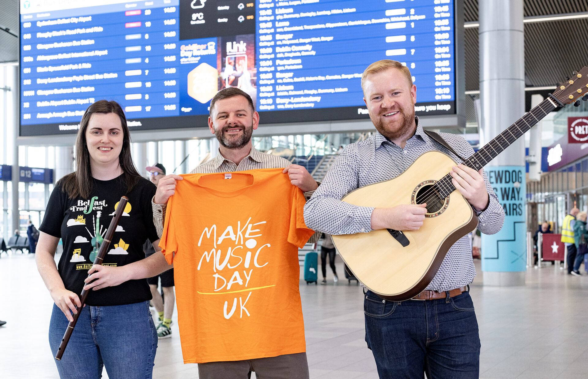Two musicians, one female and one male, pose in a station holding a flute and a guitar, while another man stands in the middle holding an orange tshirt saying Make Music Day UK