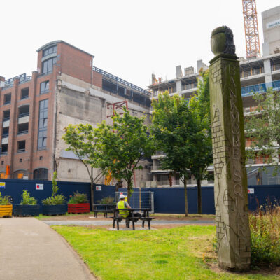 view of a public square with green grass, a large totem and blue surrounds of a building site and a multistorey car park