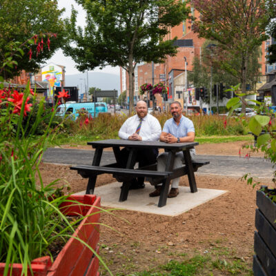 Two men in shirts sit on a bench surrounded by plants and trees