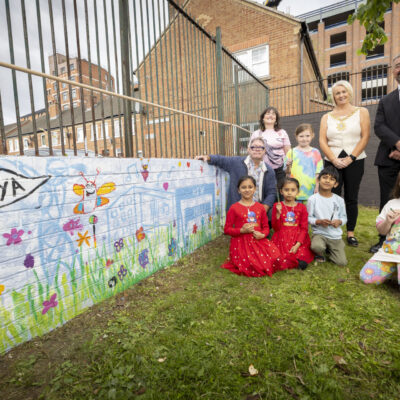 Children kneel in front of a colourful mural they have painted, with adults and the lord mayor wearing her mayoral necklace behind them. The mural is a colourful green and blue with flowers and beflast landmarks